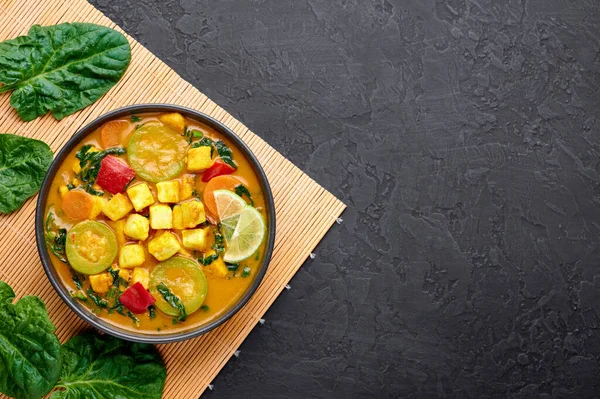 Yellow Veg Thai Curry with Tofu and vegetables in black bowl at dark slate background. Vegetarian Thai Curry with tofu, zucchini, pepper, spinach, carrot. Thai Food. Indian vegetable curry. Copy space