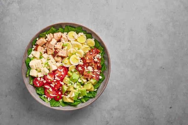 Cobb Salad in gray matte bowl on concrete backdrop. American cuisine dish with tomatoes, chicken, bacon, eggs, cheese, avocado and romaine lettuce. USA food and meal