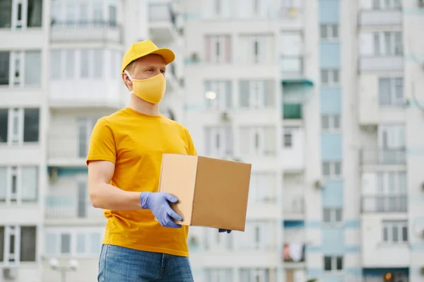 Delivery Man worker in yellow uniform cap, t-shirt, face mask and gloves holds a cardboard box package on building backdrop. Safety delivery quarantine service in covid-19 virus pandemic period.