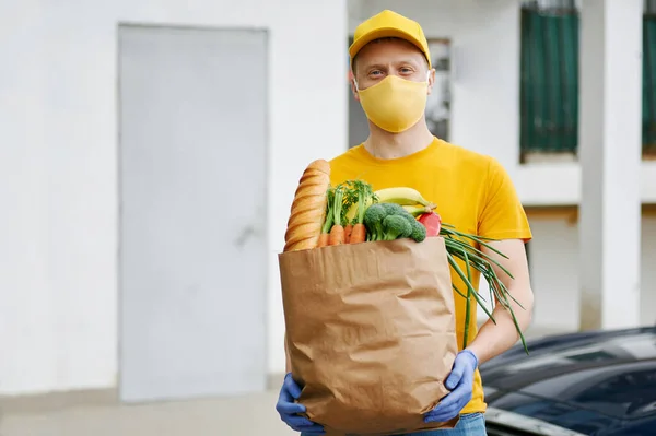 Grocery Delivery Man in yellow uniform cap, t-shirt, face mask, gloves holds a paper bag with food, fruit, vegetables on building backdrop. Quarantine delivery service in covid-19 virus pandemic.