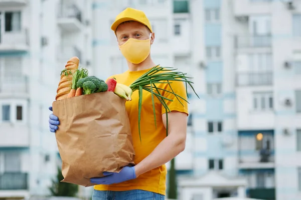 Grocery Delivery Man in yellow uniform cap, t-shirt, face mask, gloves holds a paper bag with food, fruit, vegetables on building backdrop. Quarantine delivery service in covid-19 virus pandemic.