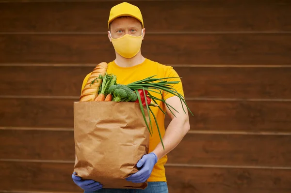 Grocery Delivery Man in yellow uniform cap, t-shirt, face mask, gloves holds a paper bag with food, fruit, vegetables on wooden backdrop. Quarantine delivery service in covid-19 virus pandemic.