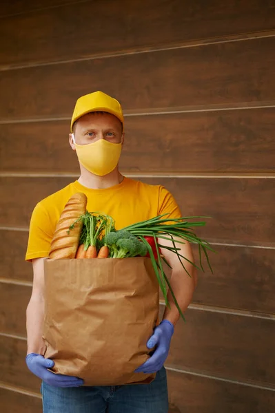 Grocery Delivery Man in yellow uniform cap, t-shirt, face mask, gloves holds a paper bag with food, fruit, vegetables on wooden backdrop. Quarantine delivery service in covid-19 virus pandemic.