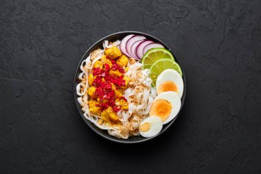 Nan Gyi Thoke in black bowl at dark slate background. Nan gyi Thohk is popular burmese cuisine dish with rice noodles, chicken breasts with spices, eggs, peanuts, lime and red onion. Top view. clipart