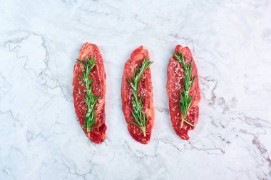 raw marbled beef steaks with green rosemary branch and sea salt at white marble background. Copy space
