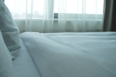 Pencereye dayalı beyaz yatak. Çerçevede yastıklar ve battaniyenin bir parçası var. Tipik otel içi.