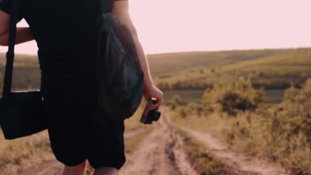 Молодий турист прогулюється по сільській дорозі посеред гори, тримаючи в руці ретро-камеру. Він закоханий у захід сонця — стокове відео