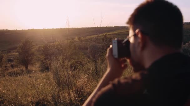 Retrato de um jovem fotógrafo com uma câmera retrocedida por trás. Jovem turista tira fotos na natureza. Admire a beleza do pôr do sol — Vídeo de Stock
