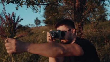 Karizmatik bir genç adam elinde bir buket kır çiçeğiyle çimlerin üzerinde oturur ve dergi için eski bir kamerayla fotoğraf çeker..
