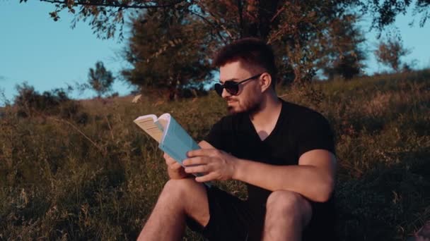 De charismatische jongeman zat in het gras op het gras en voelde zich gelukkig met het lezen van een boek. Een boek lezen midden in de natuur. Spirituele ontspanning. Vrijetijdsconcept lezen persoonlijk — Stockvideo