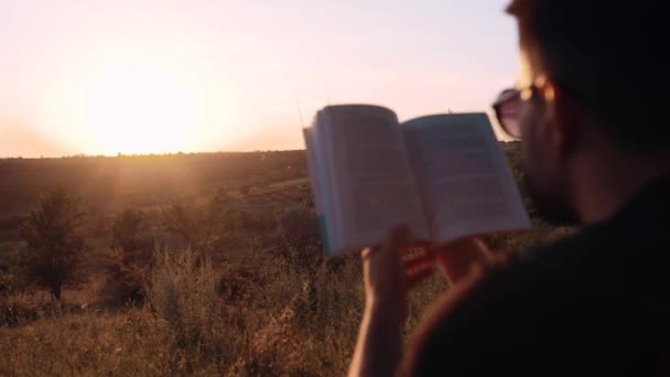 Rama boczna z tyłu z charyzmatycznym brodatym młodzieńcem czytającym książkę na zewnątrz. Cudowny krajobraz o zachodzie słońca. Relaks w sercu natury — Wideo stockowe