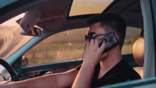 Ein brünetter Mann mit Sonnenbrille telefoniert am Steuer eines Luxusautos mit seinem Handy. Der junge Mann ist mit heller Sommerkleidung bekleidet. Blick von links auf den Fahrer — Stockvideo