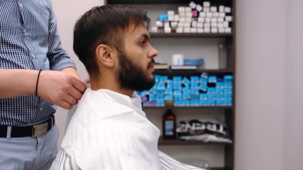Barberaren förbereder klienten för en frisyr, binder en servett. Modernt småföretagskoncept — Stockvideo