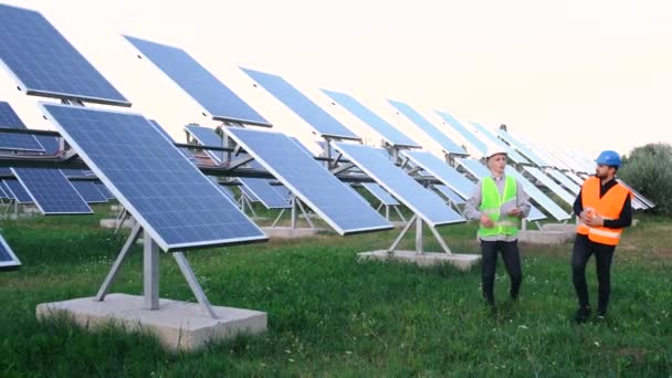 Dois engenheiros profissionais em equipamentos de proteção andando em campo com painéis solares. — Vídeo de Stock