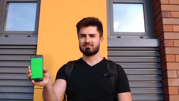 Beau jeune homme caucasien tient un téléphone à écran vert dans sa main et sourit. Un mur multicolore en arrière-plan — Video