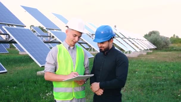 Boldog fiatal mérnökök beszélgetnek egy mező közepén napelemekkel. Megújuló energia koncepciója. — Stock videók