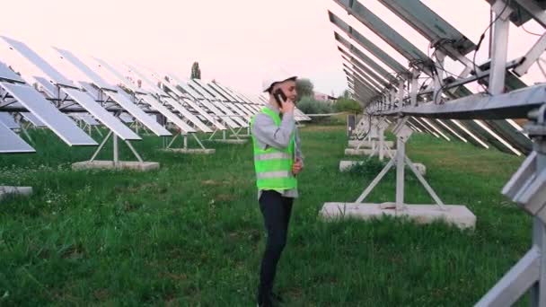 Šťastný mladý inženýr, oblečený ve speciálním vybavení, mluví po telefonu v pozadí na solárních panelech. — Stock video