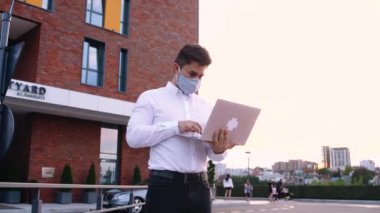 Yüz maskeli genç işadamı şehirde bankta çalışıyor, dizüstü bilgisayar kullanıyor.