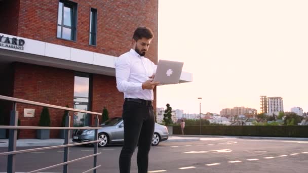 Vue latérale de l'homme barbu heureux en costume formel debout à l'extérieur avec ordinateur portable ouvert. — Video