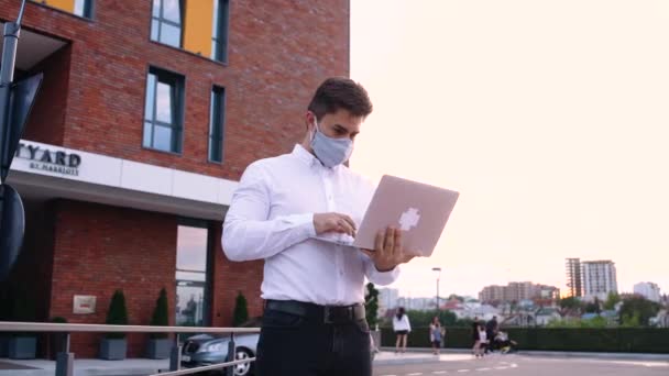 Joven hombre de negocios con máscara facial que trabaja al aire libre en el banco de la ciudad, utilizando el ordenador portátil — Vídeos de Stock