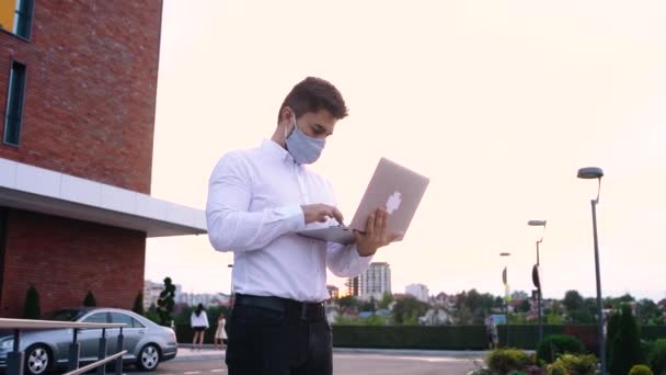 Homme d'affaires dans les masques médicaux est assis sur la terrasse d'un café et de travailler sur un ordinateur portable, vue à travers le verre. — Video