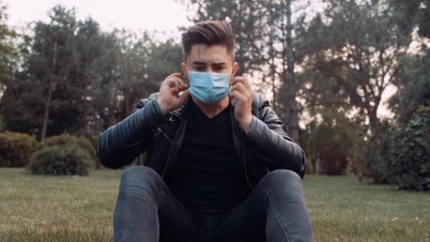 A man is sitting on the grass in the park. On his face he has a surgical mask — Stock Video