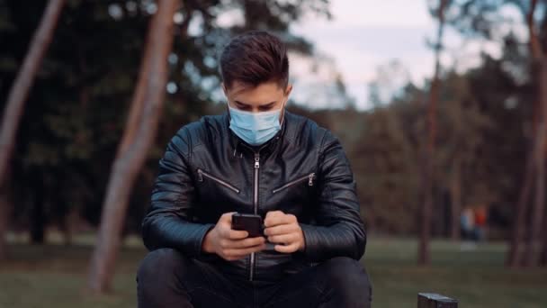 Bij zonlicht jong gelukkig man met chirurgische masker staan in het park centrum maakt gebruik van telefoon sms 'en scrollen afluisteren, — Stockvideo