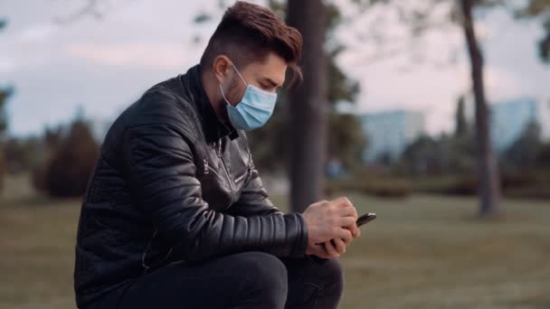 Een knappe man met een chirurgisch masker zit op de bank in het park. — Stockvideo