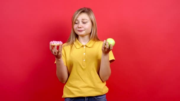 Chica bonita y gorda con una camiseta amarilla sosteniendo una dona en una mano y una manzana en otra. — Vídeo de stock