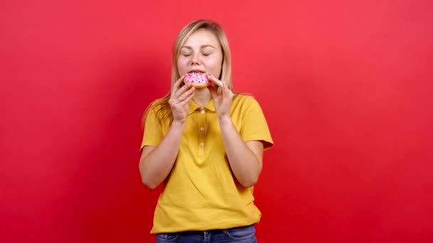 Linda chica gorda con una camiseta amarilla sosteniendo un donut en su mano, ella no puede ayudarse a sí misma y comienza a comerlo. — Vídeos de Stock