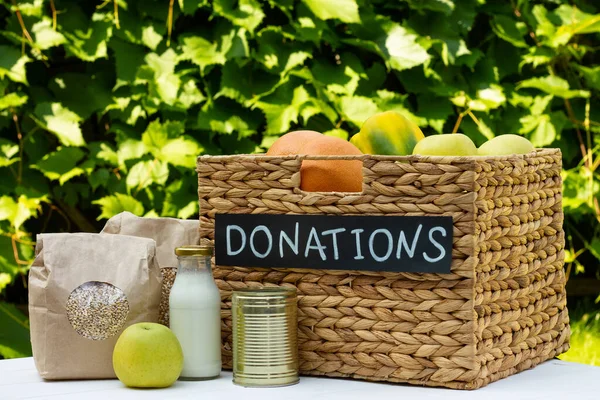 Olika livsmedel i en låda för donationer på ett bord på en bakgrund av greener. Välgörenhet och donationskoncept, kopieringsutrymme. — Stockfoto