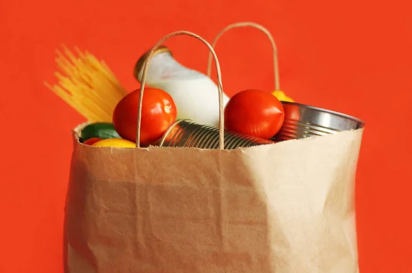 Products in a paper bag on a red background. Food in a food delivery service package. Canned food, pasta, vegetables, fruits, milk. Contactless delivery.