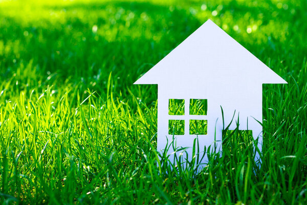 Белый силуэт игрушечного домика, вырезанного из картона на фоне травы. Маленький домик из белой бумаги. Копирование пространства.