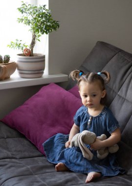 Küçük tatlı üzgün kız 1-3 kanepede peluş bir tavşanla oturuyor.