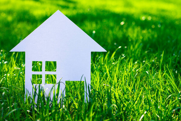 Белый силуэт игрушечного домика, вырезанного из картона на фоне травы. Маленький домик из белой бумаги. Копирование пространства.