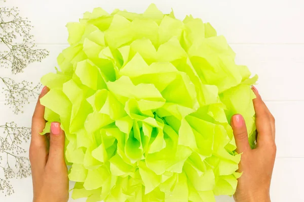 Step by step tutorial on making a light green fluffy pompom from tissue paper on a white wooden table background. Top view. Step 10