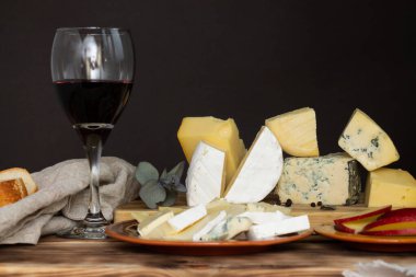 Koyu bir arka planda tahta kesme tahtasındaki farklı peynir türleri elma ve bir bardak şarapla. Boşluğu kopyala.