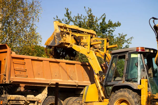 Geniş metal kazıcı kovası kum ya da kil kazar ve inşaat alanındaki bir çöp kamyonuna yükler..