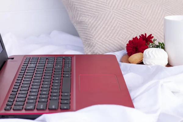 Keyboard of a red laptop, standing on the bed next to a cup, marshmallow, cookies and flowers. Online shopping, teleworking or distance learning concept