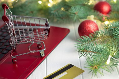 Kredi kartının yanında kırmızı bir dizüstü bilgisayarın klavyesinde alışveriş yapmak. Ampulleri ve beyaz arka planda parlak topları olan bir Noel ağacının dalları. Noel ve Yeni Yıl çevrimiçi alışveriş kavramı