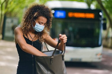 Coronavirus 'a karşı maske takan genç bir kadın otobüs durağında çantasına bakıyor.