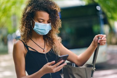 Genç bir kadın telefonunu kullanıyor ve salgın sırasında koruyucu bir maske takıyor.