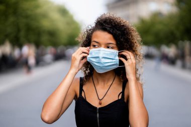 Sokakta dikilen ve maske takan bir kadın var.