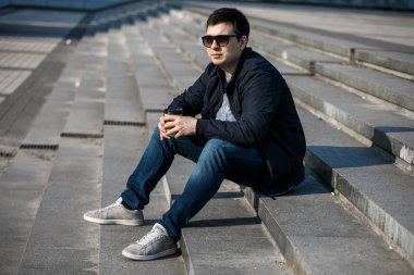 Genç bir adam şehirde kahve içer ve dışarıda yürür..