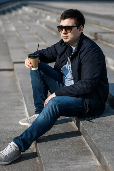 A young man drinks coffee in the city and walks outside.