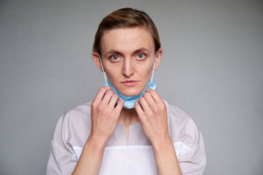 Gri arka planda koruyucu maske takan kadın doktor ya da bilim adamına yakın durun. Tıbbi maskeyi elleriyle ayarlıyor.