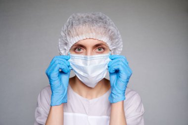 Gri arka planda tıbbi maskesi ve cerrahi kapağı olan kadın doktor ya da bilim adamına yakın durun. Elleri lateks eldivenli maskeyi ayarlıyor.
