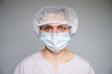 Tıbbi maskeli, cerrahi şapkalı ve gri arka planda gözlüklü kadın doktor ya da bilim adamına yakın durun..