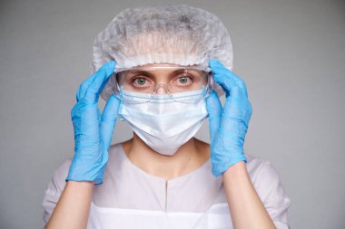 Tıbbi maskeli, cerrahi şapkalı, gözlüklü ve eldivenli kadın doktor ve bilim adamlarına yakın plan gri arka plan.