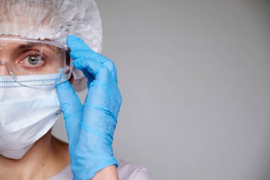 Tıbbi maskeli, cerrahi şapkalı, gözlüklü ve eldivenli kadın doktor ve bilim adamlarına yakın plan gri arka plan.